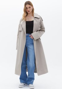 Trench coat beige chiaro con ampi revers, design con cintura e tasche laterali, abbinato a jeans a zampa d'elefante blu e sneakers bianche.