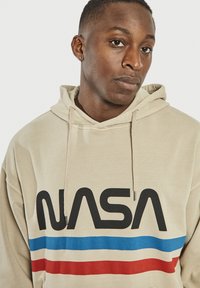 Beżowa bluza z kapturem z czarnym logo "NASA". Posiada niebieskie i czerwone poziome paski. Wykonana z miękkiego materiału, o luźnym kroju i z kapturem ze sznurkiem.