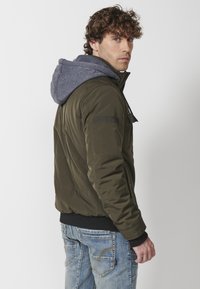 Giacca verde oliva con cappuccio grigio, dotata di chiusura a zip, polsini a costine e branding discreto sulla manica. Indossata sopra jeans blu.