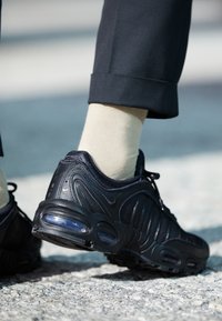 Černá atletická bota s texturovaným designem, výrazným vzduchovým polstrováním a logem, doplněná světle zbarvenými ponožkami a tmavými kalhotami.