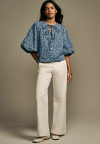 Modra cvetličasta bluzica z nabranimi rokavi, podrobnostjo v obliki ključavnice, v kombinaciji z belimi širokimi hlačami in svetlimi čevlji. Gladka tkanina in sproščen kroj.