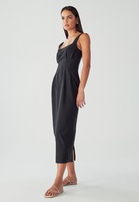 Črna midi obleka, izdelana iz gladkega materiala, z kvadratnim izrezom, širokimi naramnicami in stranskimi razporki na robu za elegantno podobo.