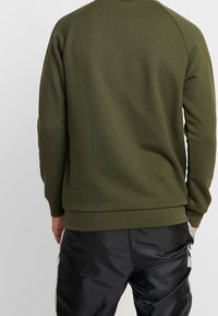 Olivgrön sweatshirt med ribbad nederkant och ärmslut, med rund halsringning och raglanärmar, i kombination med svarta glänsande byxor.