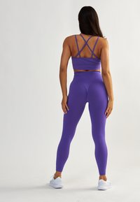 Soutien-gorge de sport violet avec des bretelles croisées et un legging taille haute assorti, confectionné dans un matériau élastique, offrant une texture lisse. Baskets blanches.