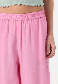 Pantalon léger rose avec une finition texturée. Il possède une taille élastique et des poches latérales, offrant une coupe décontractée.