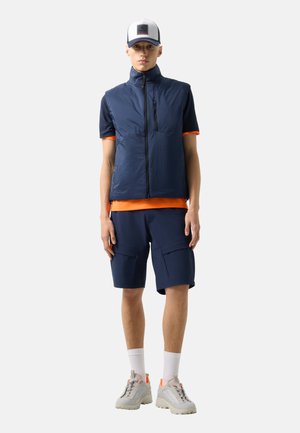 Jonge man met een marineblauw vest over een oranje shirt, marineblauwe korte broek, witte sokken, grijze sneakers en een witte baseballpet.