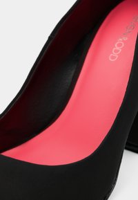 Chaussure à talon noir avec finition lisse et semelle intérieure rose vif présentant un texte de marque partiel sur fond blanc.