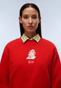 Κόκκινο φούτερ με γραφικό σχέδιο ενός κουνελιού με γυαλιά ηλίου και ποτήρι, με γιακά από πράσινο και λευκό γκίνγχαμ από κάτω.