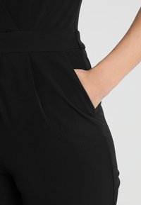 Main droite placée dans la poche latérale d’un pantalon noir ajusté sur un fond clair uni.
