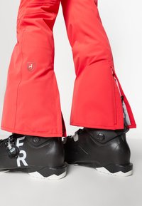 Crvene ski hlače s lepršavim dizajnom, koje imaju patentne zatvarače na gležnju. Nosene su preko crnih ski čizama s bijelim natpisima i teksturiranom finishem.