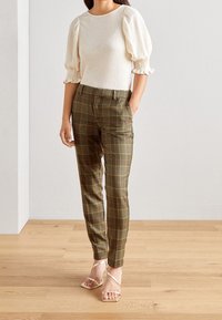 Top con maniche a palloncino e polsini arricciati abbinato a pantaloni attillati a quadri verde oliva con linee rosa e gialle. Sandali dorati con cinturini.
