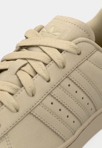 Beige athletic shoe with a smooth suede-like finish, featuring three parallel stripes, textured laces, and a subtle logo on the tongue.