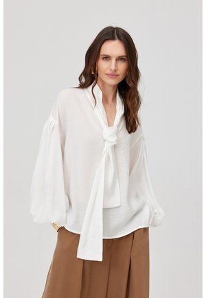 Kobieta o długich falowanych włosach, ubrana w białą bluzkę z dużym zawiązanym krawatem i obszernymi rękawami oraz brązowe, wysokie, szerokie spodnie.