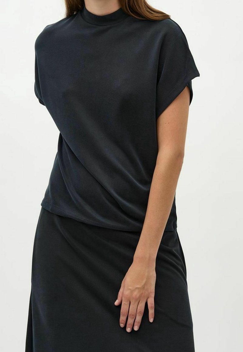 Femme portant un haut noir à manches courtes avec un col montant et une jupe noire assortie, debout avec une main reposant sur sa cuisse.