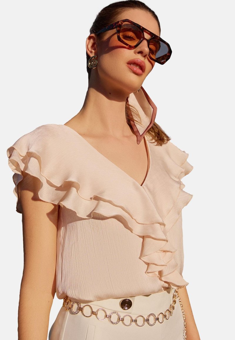 Il modello posa con un'espressione sicura, indossando una blusa ruffled rosa, occhiali da sole oversize e un orecchino di design, su uno sfondo bianco.