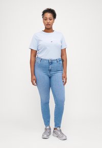 Camiseta de manga corta azul claro con un logotipo bordado, combinada con unos jeans ajustados azul claro y zapatillas deportivas plateadas. Fondo blanco liso.
