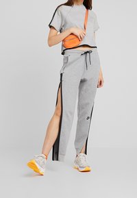 Šedé krátké tričko a šedé teplákové kalhoty se zipy na bocích, s černými detaily. Obsahuje oranžovou křížovou tašku a světlé tenisky.