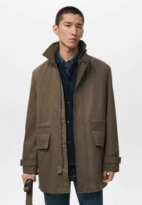 Chaqueta impermeable marrón con un cuello alto, botones a presión en el frente, dos grandes bolsillos frontales y puños ajustables, llevada sobre una camisa oscura.