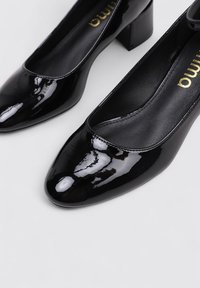 Chaussures à talon bloc en cuir verni noir avec un bout arrondi et un design épuré. Texture lisse avec une finition brillante et une marque interne.