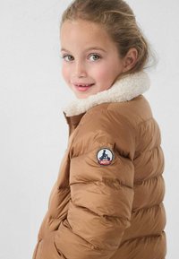 Chaqueta acolchada marrón tipo puffer con diseño acolchado y cuello de piel sintética blanca. Cuenta con un parche de logo redondo en la manga izquierda.