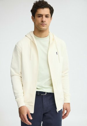 Hombre con sudadera con cremallera color crema sobre una camisa verde claro y pantalones azul marino contra un fondo claro y sencillo.