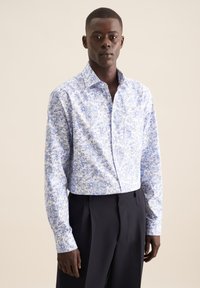 Camicia bianca con motivo floreale e geometrico blu, colletto a punte, chiusura con bottoni frontale e maniche lunghe, abbinata a pantaloni neri su misura.