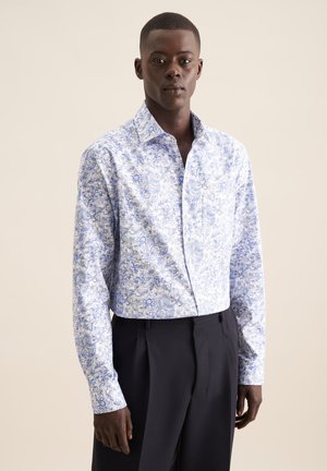 Witte blouse met een blauw bloemen- en geometrisch patroon, met een gespreide kraag, knoopsluiting aan de voorkant en lange mouwen, gecombineerd met zwarte getailleerde broeken.