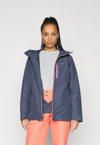 Αδιάβροχο ναυτικό μπουφάν με κουκούλα. Διαθέτει ροζ λεπτομέρειες, τσέπες με φερμουάρ και ρυθμιζόμενες μανσέτες. Φοράει πάνω από έναν γκρι τοπ και πορτοκαλί παντελόνι.