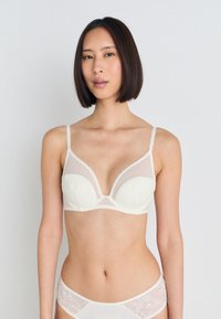 Cream-colored bra with sheer mesh cups, a deep V-neckline, and adjustable straps. Lace accents on the sides add texture.