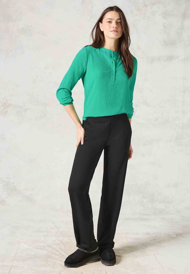 Grön plisserad skjorta med knappar och trekvartsärmar, i kombination med svarta vida byxor och mörka mockaskor med dekorationer.