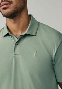 Polo de color verde claro hecho de tela texturizada con una tapeta de tres botones y un logo de ciervo bordado en el lado izquierdo del pecho.