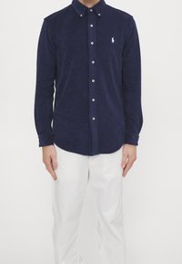 Mörkblå skjorta med knappar i mjukt tyg med en texturerad yta, försedd med en klassisk krage och en liten vit logotyp på bröstet.