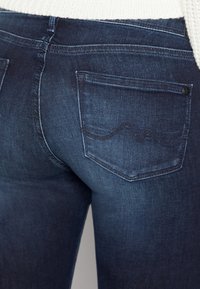 Tmavé džíny z denimu s přiléhavým střihem. Obsahují zadní kapsy s jemným detailem prošití. Materiál má hladkou texturu a mírný lesk.