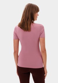 Úzký top s krátkým rukávem v světle růžové barvě, vyrobený z hladkého, elastického materiálu. Pohled zezadu ukazuje zaoblený výstřih a jednoduchý design.