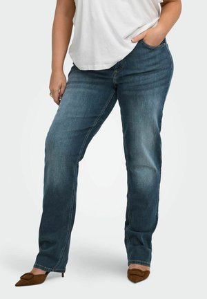 Person iført blå straight-leg jeans, hvid top og brune spidse hæle med den ene hånd i lommen.