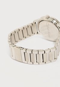 Ασημένιο βραχιόλι με μεταλλικούς κρίκους, με την επωνυμία Calvin Klein στο κούμπωμα και στην πίσω πλευρά του ρολογιού, στρογγυλή κάσα και κορώνα ρύθμισης, σε λευκό φόντο.