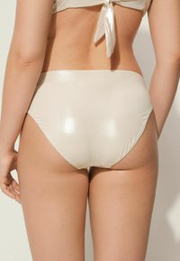 Bikiny s vysokým pasem v lesklé, světle béžové tkanině. Vyznačuje se hladkým povrchem a obrysovým tvarem s minimálním zakrytím.