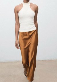 Mujer con un top blanco sin mangas tipo halter y una falda larga de seda marrón, caminando sobre un suelo y fondo de color claro y uniforme.