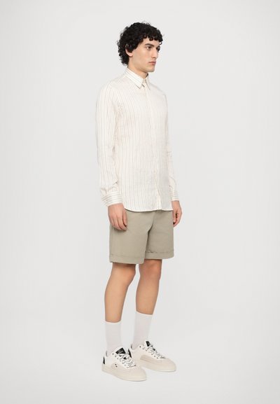 Camisa de botonadura ligera en color crema con finas rayas verticales, acompañada de pantalones cortos verdes claros y zapatillas blancas, de pie sobre un fondo neutro.