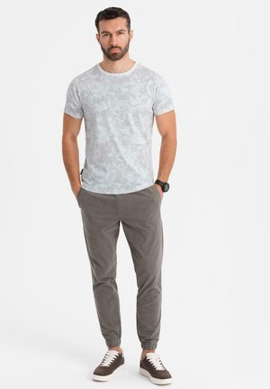 Lichtgrijze t-shirt met bloemenpatroon voor heren, gecombineerd met grijze taps toelopende broek en bruine sneakers. T-shirt heeft een ronde hals; de broek heeft elastische boorden.