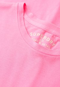 Camiseta de algodón rosa con cuello redondo. Presenta una etiqueta en texto claro y en relieve que dice "SUPERDRY CO. CRAFT & DETAIL" en el interior.