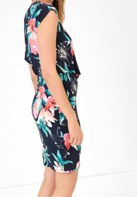 Kobieta ubrana w bezrękawową, sięgającą do kolan granatową sukienkę w różowe i zielone kwiatowe wzory, stojąca bokiem na białym tle.