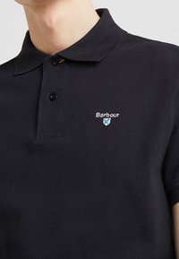 Černé polo tričko vyrobené z texturované látky, s knoflíkovou légu na dva knoflíky a vyšívaným logem "Barbour" na levé straně hrudi.