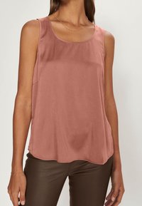 Kvinna iklädd en ärmlös blus i dammig rosafärg med rund halsringning och mörkbruna figurnära byxor, visad från axlarna till höfterna.