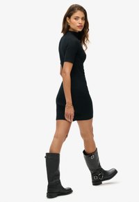 Robe noire ajustée à manches courtes, longueur genou, texture lisse. Portée avec des bottes noires montantes avec boucles. Le modèle pose de côté.