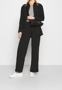 Giacca nera oversize con bottoni su un top grigio, abbinata a pantaloni larghi neri e sneakers bianche. Tiene una piccola borsa nera.