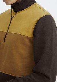 Fleece-Pullover mit hohem Kragen, der einen gelben Oberbereich und dunkelbraune Ärmel aufweist, die kontrastierende Texturen und Nähte zeigen.