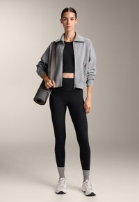 Veste gris zippée, crop top noir, leggings noirs taille haute, chaussettes grises et baskets blanches. Tiens un tapis de yoga enroulé.