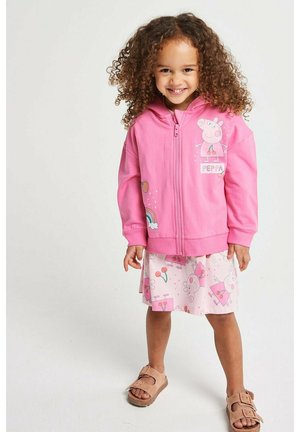 Mädchen mit lockigem Haar, das einen rosa Peppa Wutz Hoodie, passenden Rock und Sandalen trägt, lächelnd und vor einem schlichten weißen Hintergrund steht.