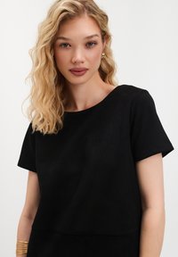 Młoda kobieta z falowanymi blond włosami, nosząca złote kolczyki, czarną bluzkę z krótkim rękawem oraz złotą bransoletkę, patrząca bezpośrednio w kamerę.